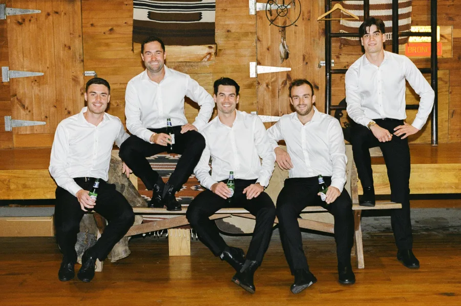 Groomsmen in white shirts and dark trousers seated together inside The Barn at Kauri Bay