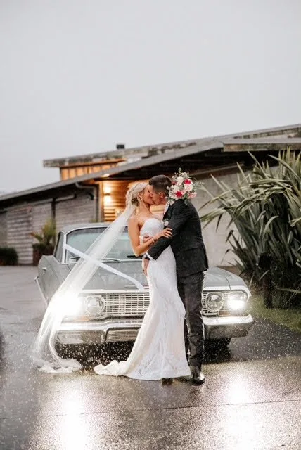 Bride and groom kissing in front of a vintage car at Kauri Bay, with the lodge behind in soft rain