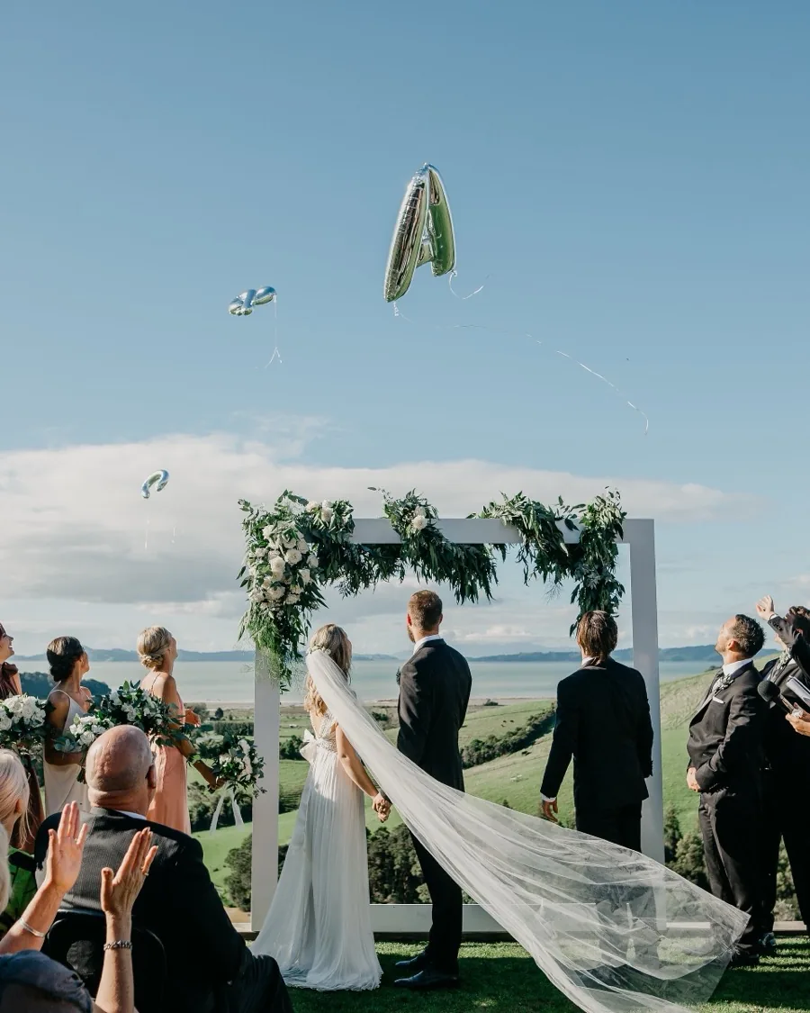Bride and groom at the floral arch on the ceremony lawn, Hauraki Gulf hills behind