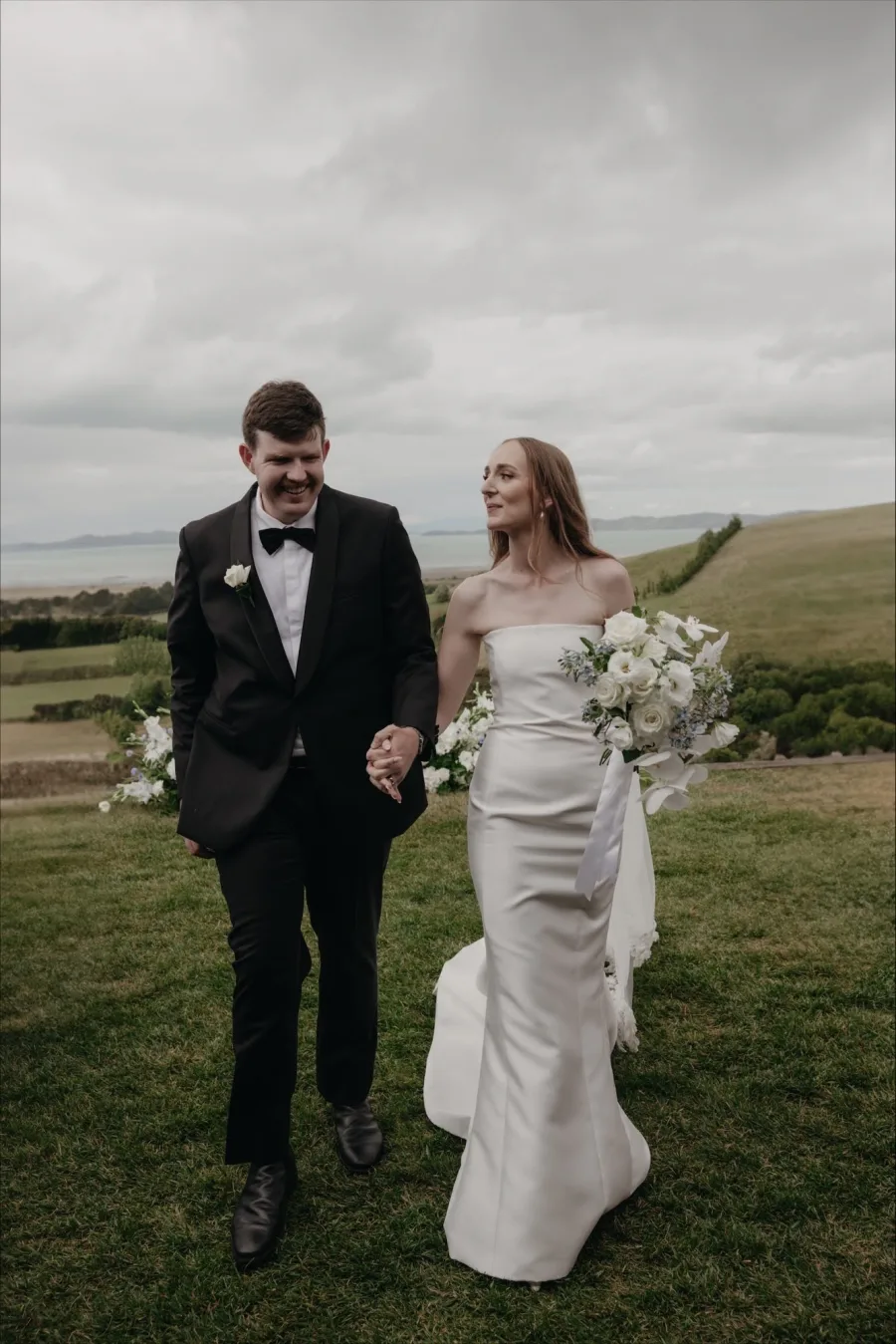 Bride and groom walking hand-in-hand on the Kauri Bay hills with the Hauraki Gulf behind