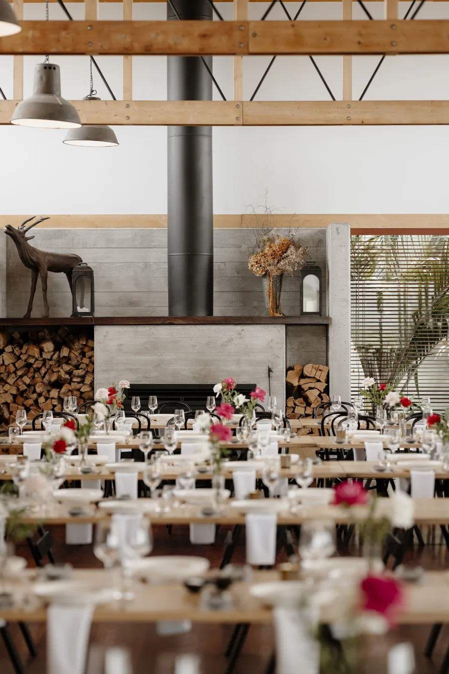 Long banquet tables set with florals and candles inside the Kauri Bay dining hall, fireplace and stacked firewood behind