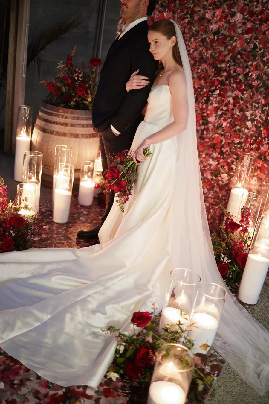 Bride and groom against a wall of red florals, surrounded by pillar candles and rose petals